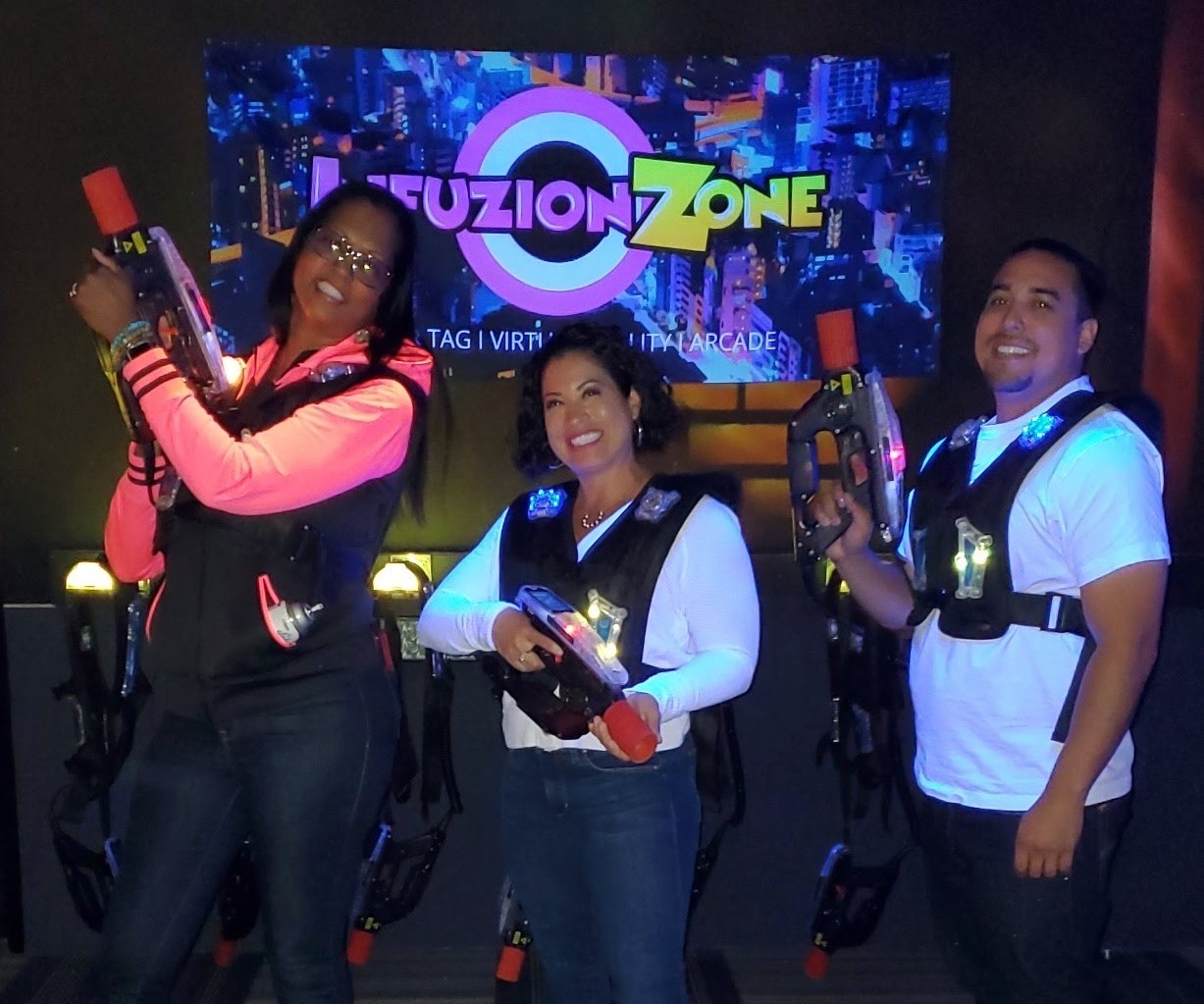 Three people smiling and posing in a laser tag arena with neon lights.